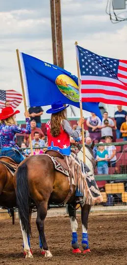 Two riders on horses carry flags in an arena while a crowd watches.