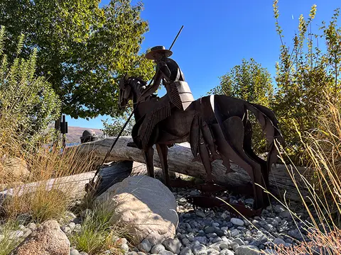 A metal sculpture of a rider on horseback stands among rocks and foliage.
