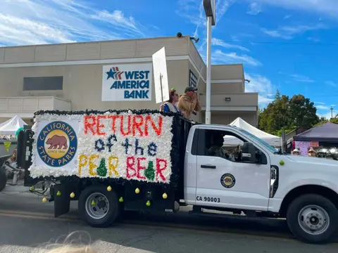A decorated parade float featuring a California State Parks sign and the words 'Return of the Pear Bear' on a white truck.