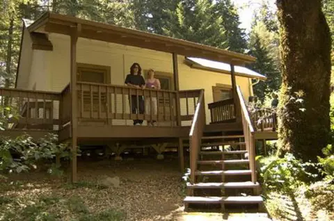 Two people standing on the porch of a cabin in the woods.