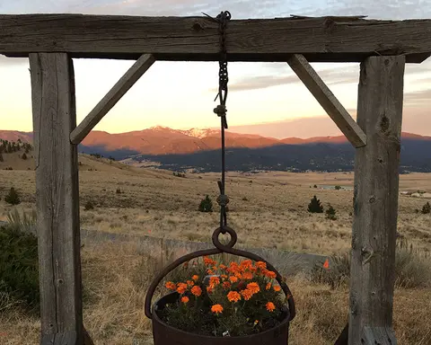 A rustic wooden frame holding a hanging pot of orange flowers with mountains in the background at sunset.