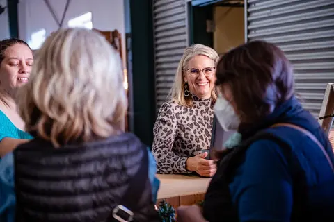 A woman smiles behind a counter while speaking with customers indoors.