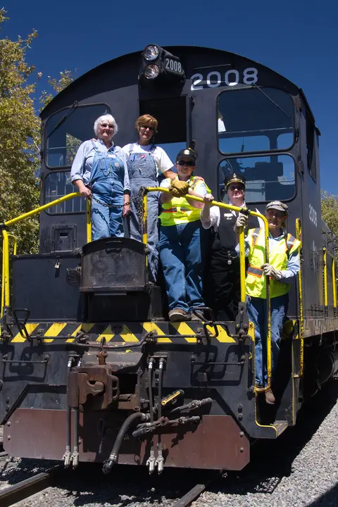 Five people pose on the front platform of a black locomotive.