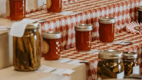 A display of various jars of preserves on steps covered with a red and white checkered tablecloth.