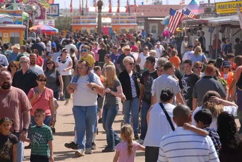 A large crowd walks along an outdoor fairground path with booths and flags.