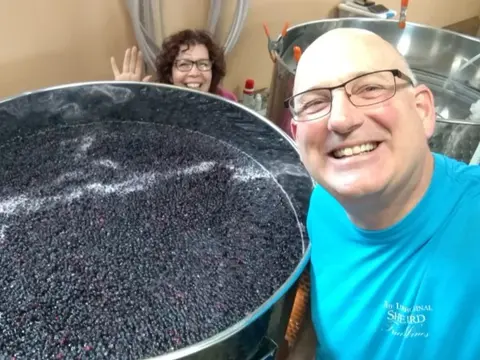Two smiling people pose inside with a large stainless steel vat filled with dark fruit.