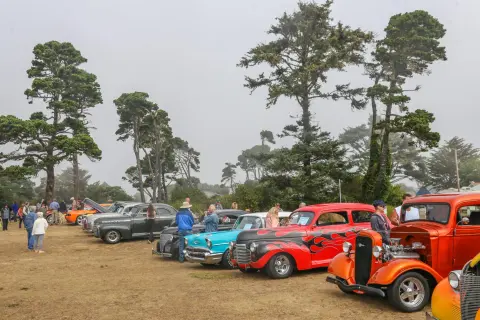 Classic cars are displayed at an outdoor gathering with people walking around them.