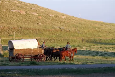 A covered wagon and riders travel across a grassy field near a hillside.