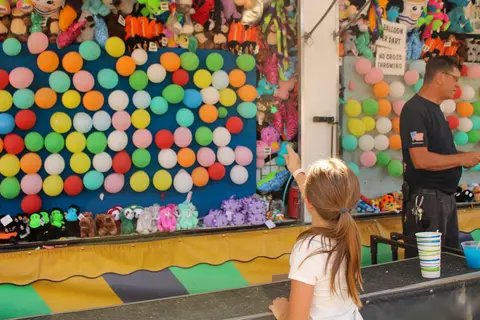 A young girl points at a carnival balloon dart game while a man stands nearby.