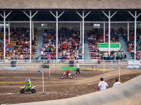 Children ride dirt bikes on a dirt track in front of a grandstand filled with spectators.