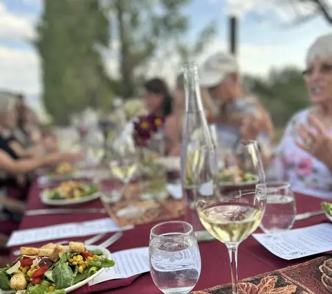People dining outdoors at a long table set with food, wine glasses, and a bottle of water.
