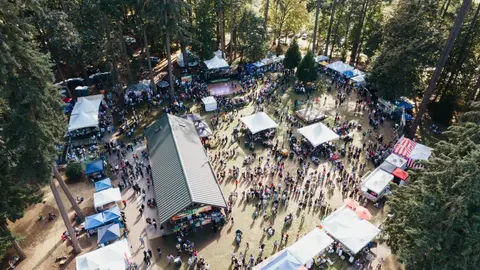 An aerial view shows a large crowd gathered among tents and trees at an outdoor event.