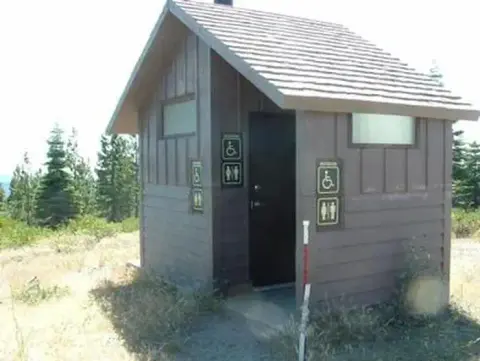 A small wooden restroom building outdoors with pine trees in the background.