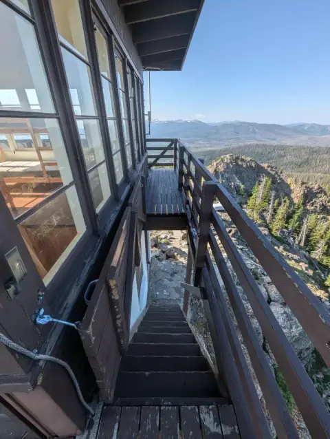 A fire lookout tower with wooden stairs and catwalk overlooking a mountain landscape.