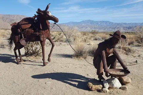 Metal sculptures of a prospector and a mule in a desert landscape.