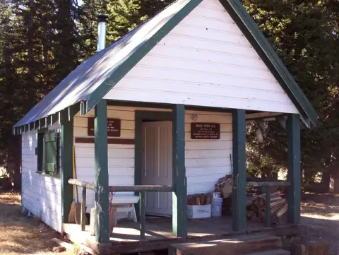 A small white wooden cabin with a covered porch sits among trees.