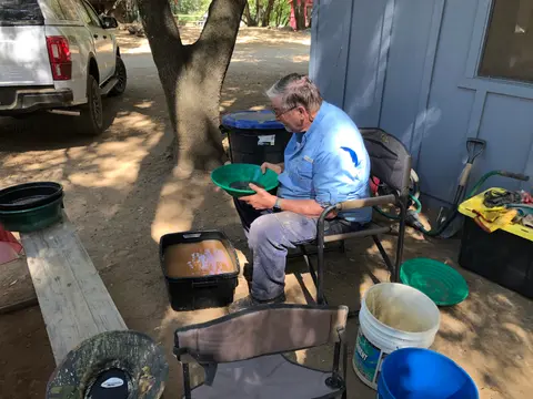 A man sits in a chair panning for gold outside next to a building and vehicle.