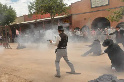 People in period clothing perform a shootout reenactment on a dusty street with spectators watching.