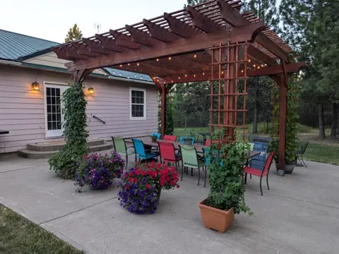 A wooden pergola with string lights is built over a patio table and chairs next to a house.
