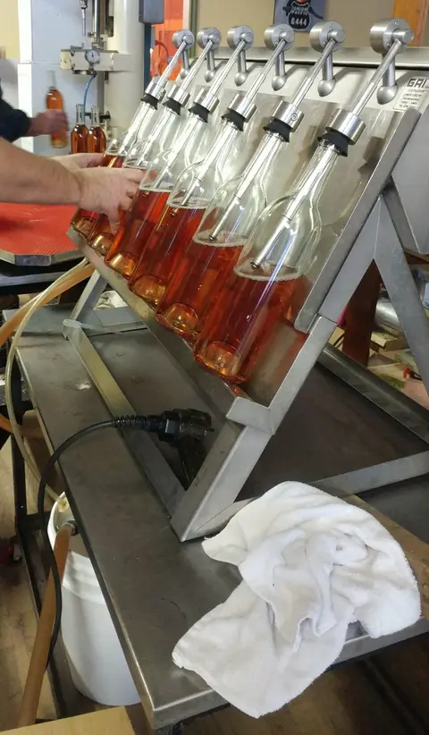 A person operates a multi-spout bottling machine filling glass bottles with amber liquid.