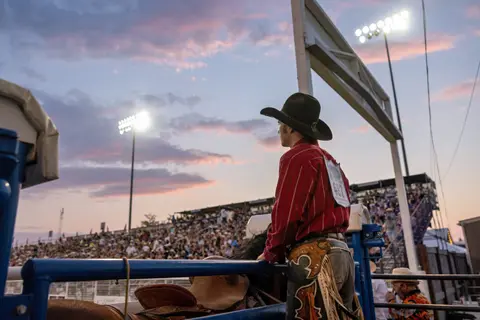 A cowboy wearing a red shirt and hat looks toward a crowded arena under a twilight sky.