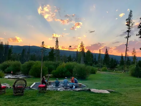 A group of people sitting around an outdoor fire pit at sunset with mountains in the background.