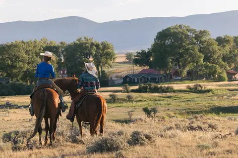 Two riders on horseback look out over a valley and mountains.