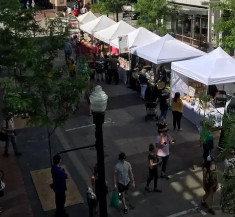 An elevated view of an outdoor market with white tents and shoppers walking along a paved walkway.