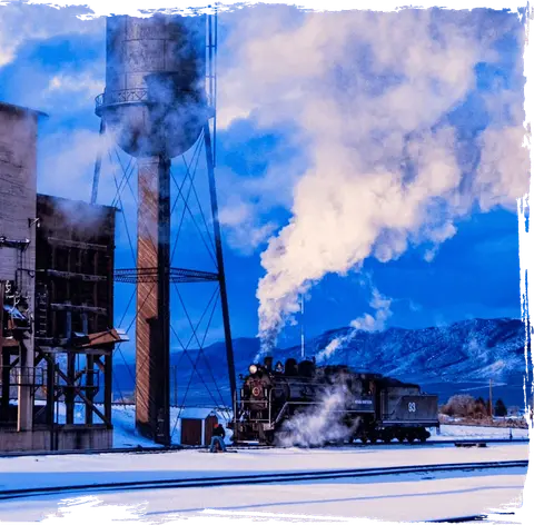 A steam train sits on snowy tracks next to an industrial water tower and mountains.