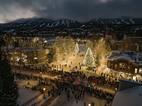 An elevated night view of a snow-covered town square decorated with glowing Christmas trees and a crowd gathered along a street.