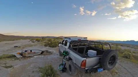 A pickup truck parked near a natural hot spring tub in a desert landscape at sunset.