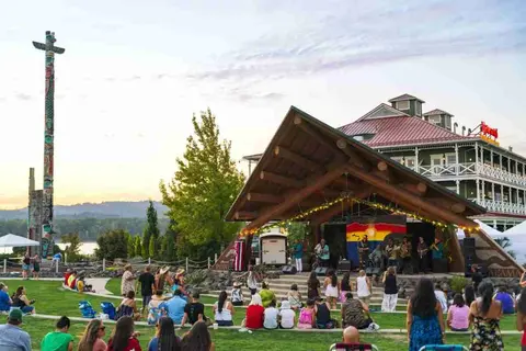 People gather on a lawn to watch a band perform on an outdoor stage near a tall totem pole and a large building.