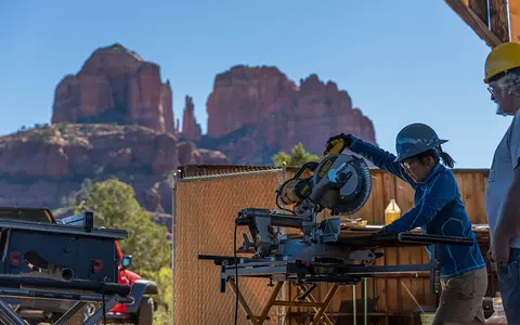 A person operating a miter saw outdoors with a large red rock formation in the background.