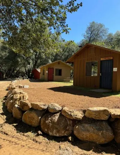 A row of rustic wooden cabins stands behind a low stone wall under trees.