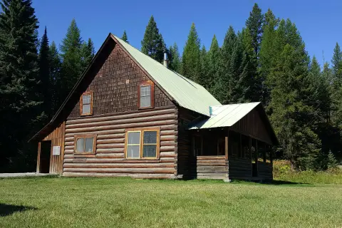 A rustic log cabin sits surrounded by tall pine trees under a clear blue sky.