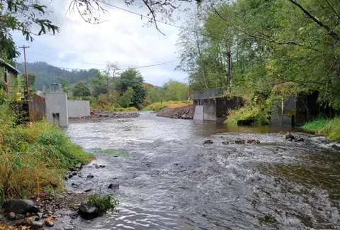 A river flows past concrete walls and green banks under a cloudy sky.