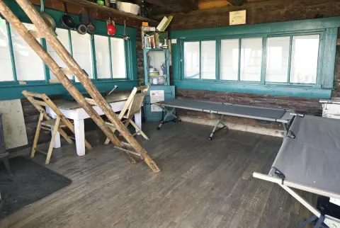 An interior view of a rustic log cabin with camping cots, windows, and cookware.