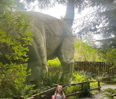 A young girl looks up in wonder at a large dinosaur statue in a wooded setting.