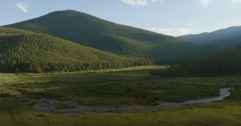 A winding river flows through a green valley surrounded by forested mountains under a blue sky.