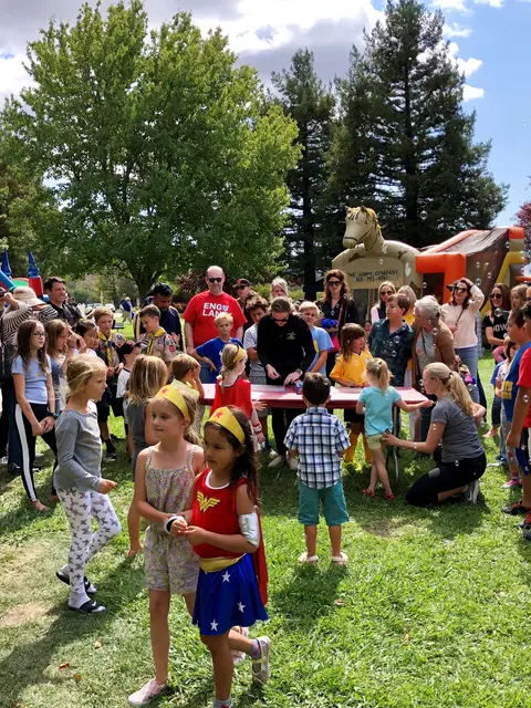 A group of children and adults gathered outdoors on grass near a bouncy house.