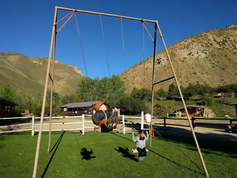 Two children swing on a large swing set outdoors with mountains in the background.