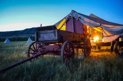 A covered wagon parked outside a lit tent in a field at dusk.