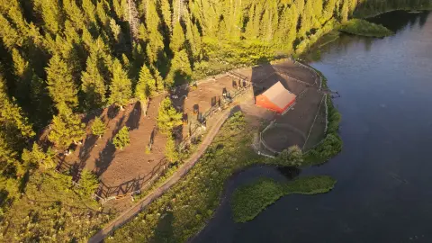 An aerial view of a red barn and corral next to a lake surrounded by forest.