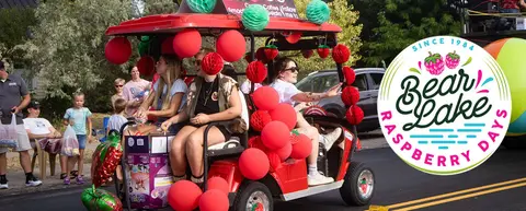 People ride in a red golf cart decorated with balloons and red honeycomb decorations during a parade.