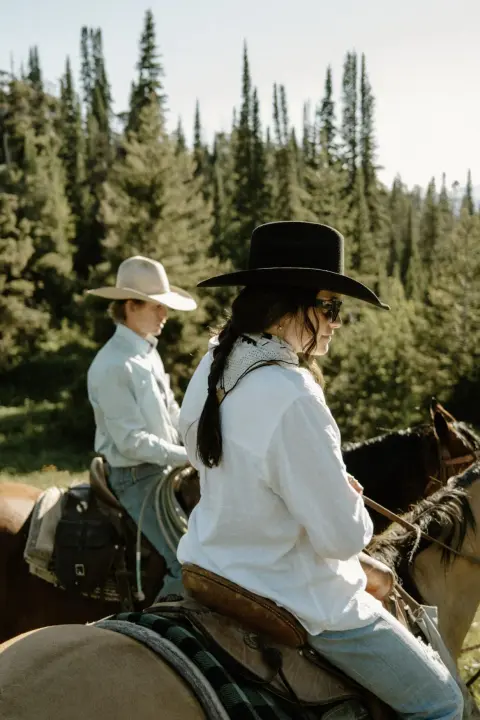 Two people ride horses through a forested area.