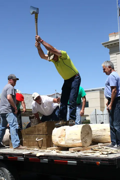 A man swings an axe into a log on a trailer while other men look on.