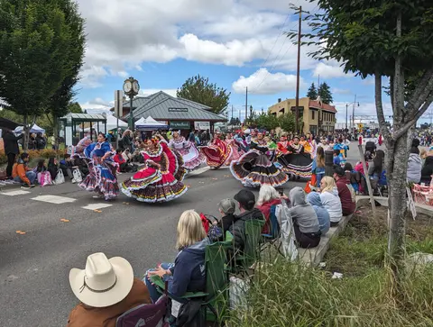 Dancers in colorful traditional dresses perform in a street parade watched by a crowd.