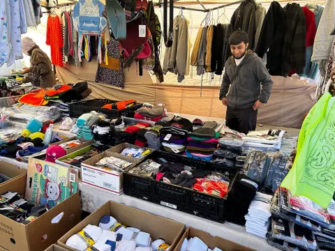 A vendor stands at a market stall filled with clothing, hats, and socks.