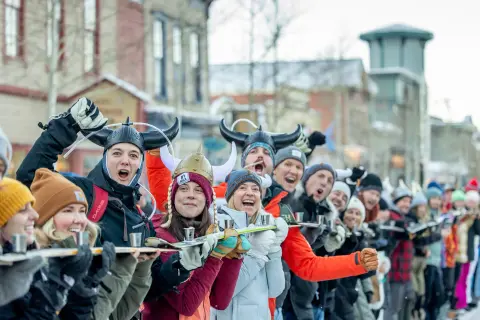 A large group of people in winter clothing and viking hats line up outdoors, holding a long ski loaded with small shots.