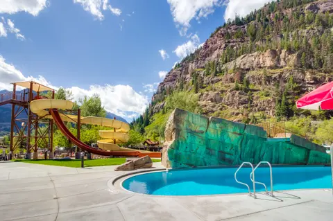An outdoor swimming pool with a water slide and a climbing wall against a mountain backdrop.